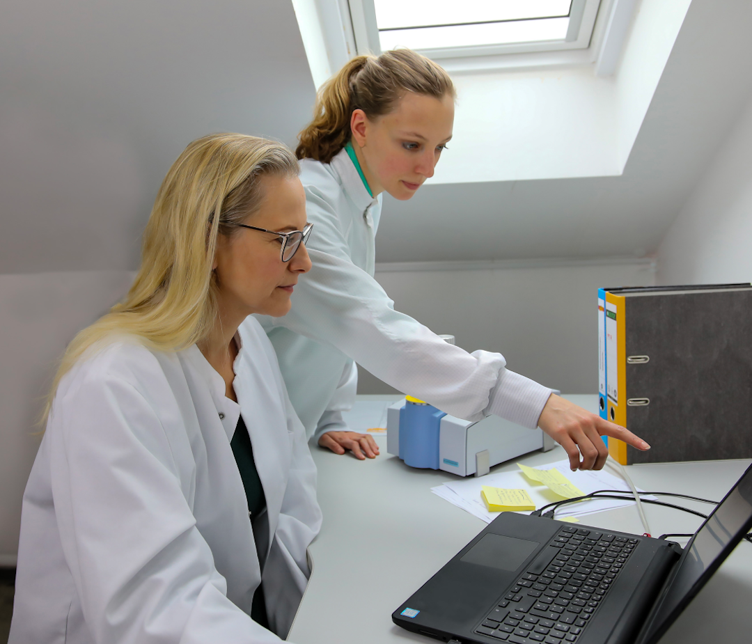 Two women in lab coats. One is pointing to a laptop screen.